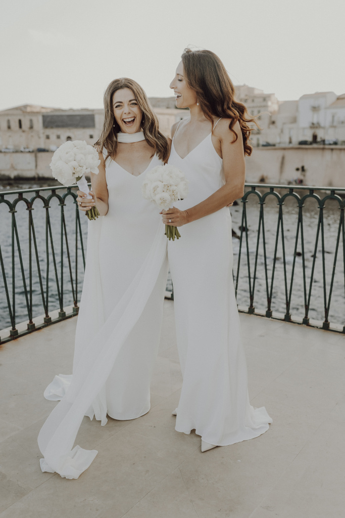 Two women in bridal gowns laughing together on baroque staircase in Ortigia Syracuse
